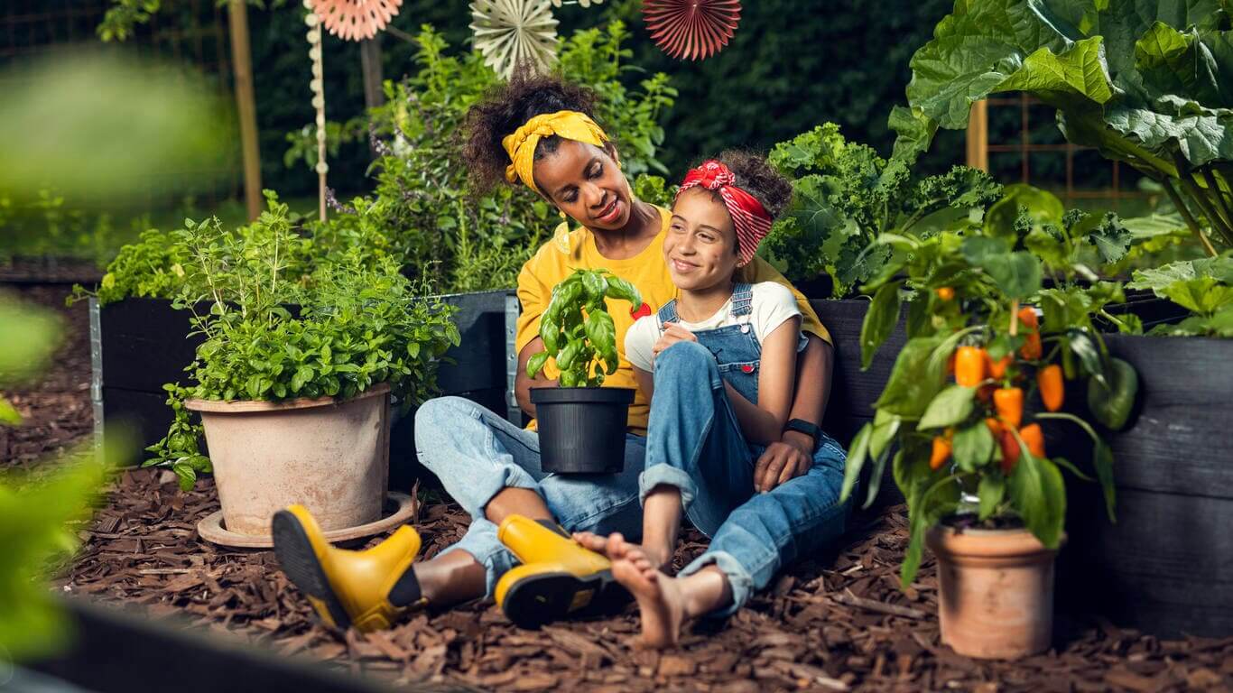 mamma och dotter sitter i trädgårdsland bland krukor och gröna odlingar