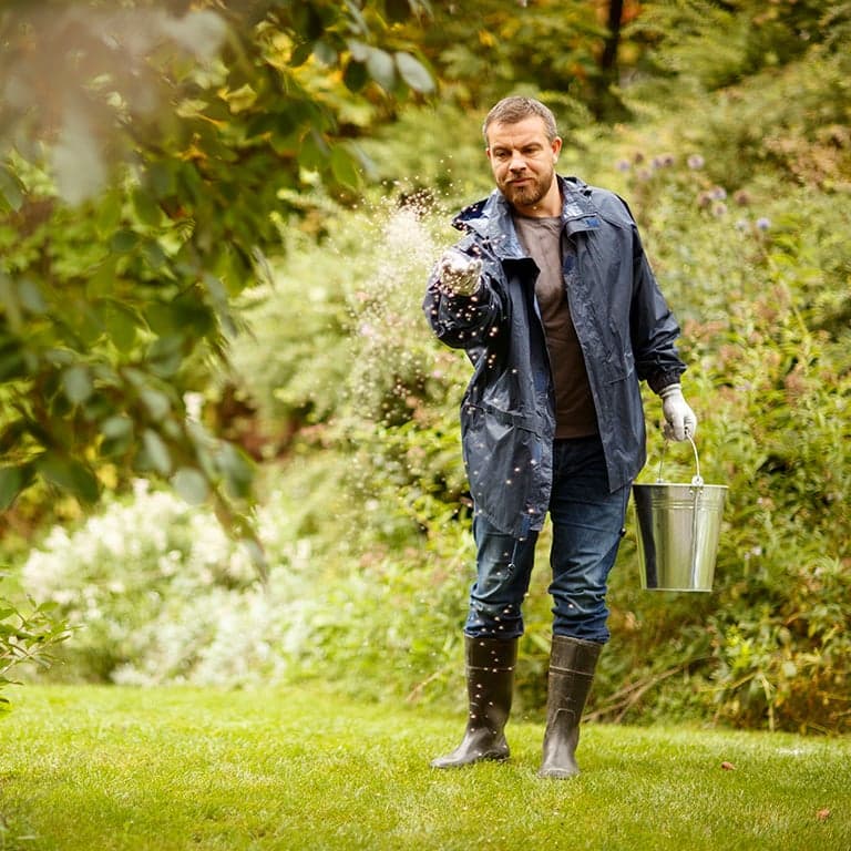 man som gödslar gräsmattan med höstgödsel i hink och gummistövlar